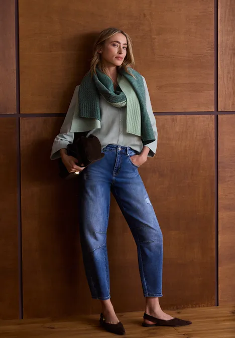 Une femme en jean, pull et châle vert appuyée sur un mur de bois, tenant un chapeau.