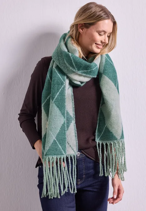Langer Schal mit Fransen und Argyle-Muster stormy green Frau vor weißer Wand trägt braunen Pullover, Jeans und langen grünen Schal mit Fransen.