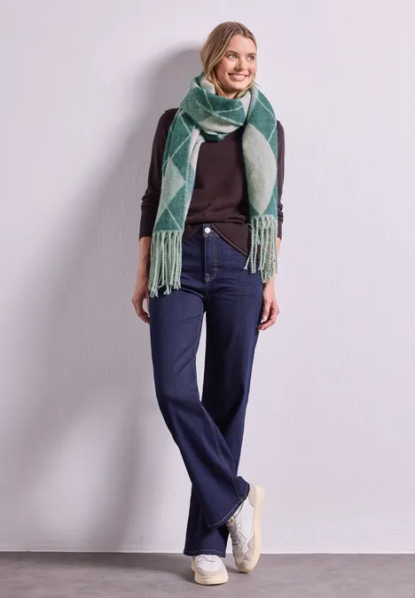 Langer Schal mit Fransen und Argyle-Muster stormy green Frau in lässigem Outfit: dunkler Pullover, weite Jeans, weiße Sneaker und grüner Schal mit Muster vor neutralem Hintergrund.