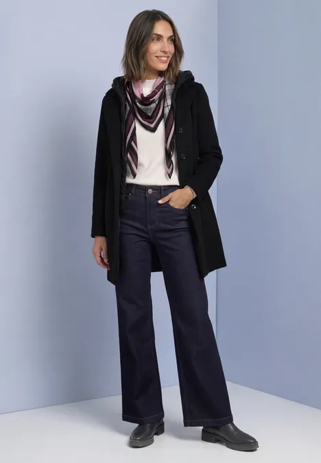 Frau in schwarzer Jacke, helles Oberteil, Schal und weiten Jeans, steht lächelnd vor hellem Hintergrund.