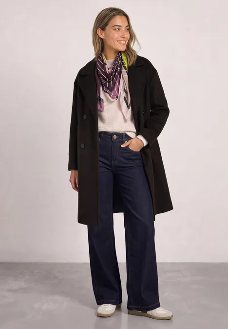 Frau in schwarzem Mantel, weißem Pullover, weiten Jeans, weißen Schuhen und gemustertem Schal.