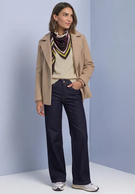 Vrouw in beige jasje, crèmekleurige trui, kleurrijk sjaaltje, wijde jeans en witte sneakers.