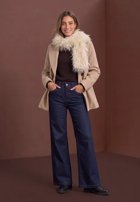 Frau in hellbraunem Mantel mit Fellkragen, braunem Pullover, hochgeschnittener Schlagjeans und schwarzen Stiefeletten.