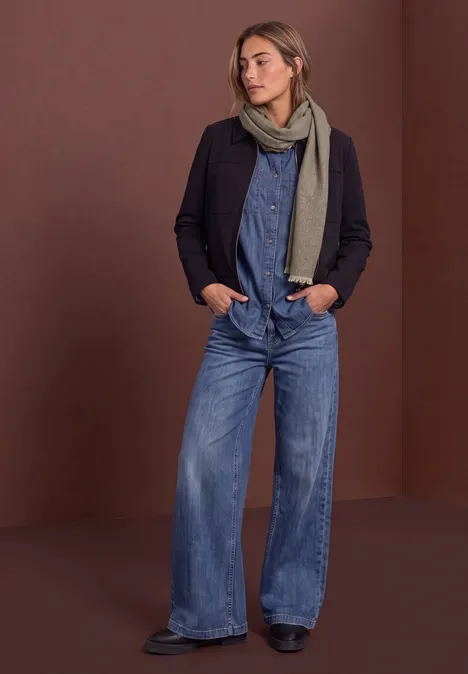 Frau in weitem Denim-Overall mit schwarzem Blazer und grauem Schal vor schlichtem Hintergrund.