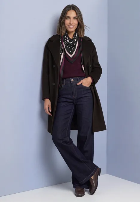 Une femme portant un manteau sombre, un haut bordeaux, un jean, une écharpe et des bottes sur fond neutre.