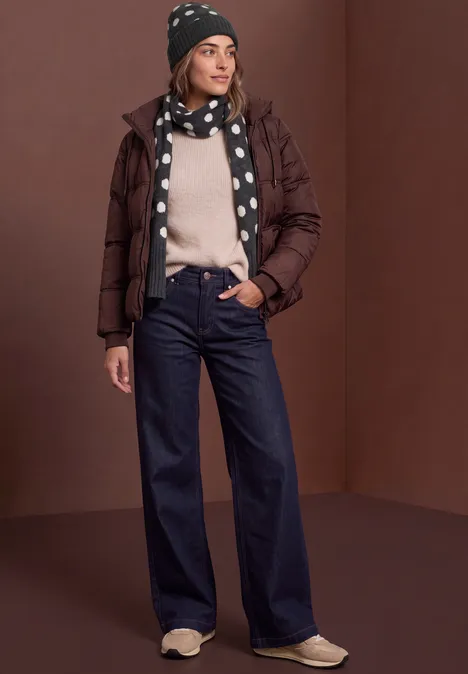 Vrouw in bruine donsjas, gestippelde sjaal en muts, lichte trui, wijde jeans en sneakers.