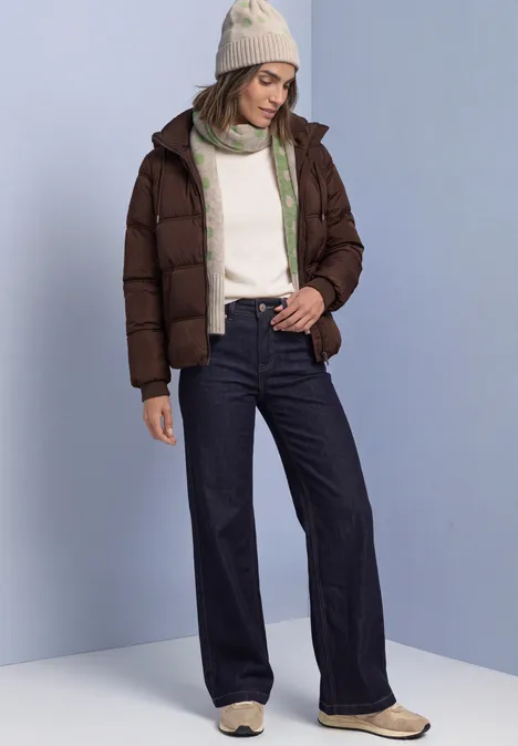 Femme en doudoune marron, pull blanc, jean large et bonnet, debout devant un fond bleu clair.