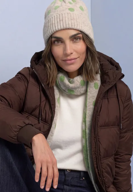 Femme en doudoune marron, bonnet crème et écharpe tricotée vert clair à motifs, adossée détendue.