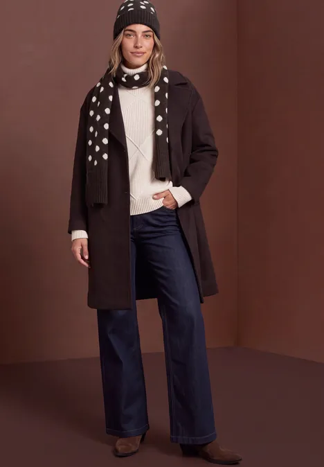 Vrouw in donkere jas, witte trui, blauwe jeans, muts en sjaal met stippen, bruine achtergrond.