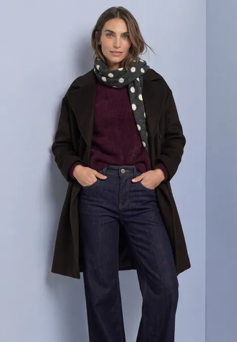Vrouw in zwarte mantel, gestippelde sjaal, bordeaux trui en donkere jeans, handen in zakken, lichte achtergrond.