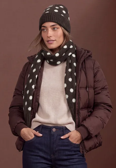 Vrouw met bruine gewatteerde jas, beige trui, blauwe jeans, gestippelde sjaal en muts.