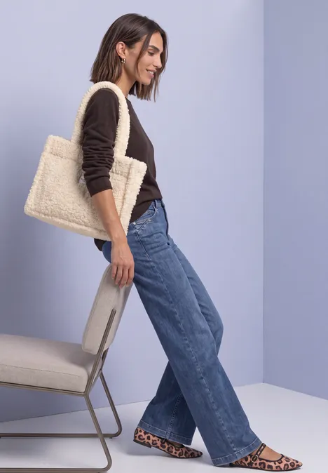 Teddy-Tragetasche in Unifarbe barley beige Frau lehnt seitlich an Stuhl, trägt dunkles Oberteil, weite Jeans, Leopardenmuster-Slipper und beige Shopper-Tasche.