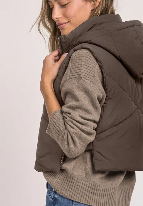 Frau in brauner Steppweste mit Kapuze über beigem Strickpullover, seitlich stehend und lächelnd.
