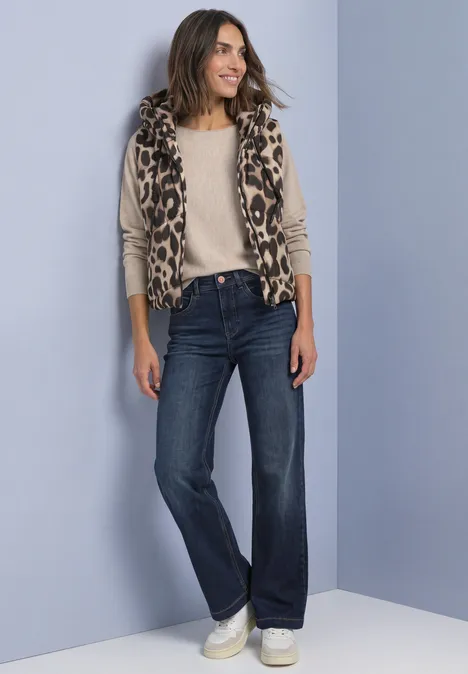 Frau in legerem Outfit: Leo-Weste, helles Shirt, weite Jeans und weiße Sneaker vor grauem Hintergrund.