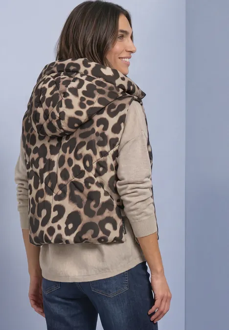 Frau von hinten in leo-bedruckter, wattierter Kapuzenweste, beigem Pullover und Jeans.