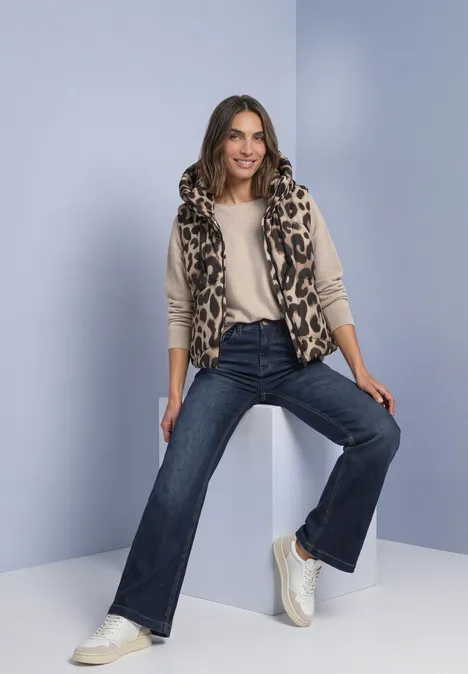 Frau in Leo-Weste, beigem Top und ausgestellten Jeans sitzt lässig auf einem weißen Block vor blauem Hintergrund.