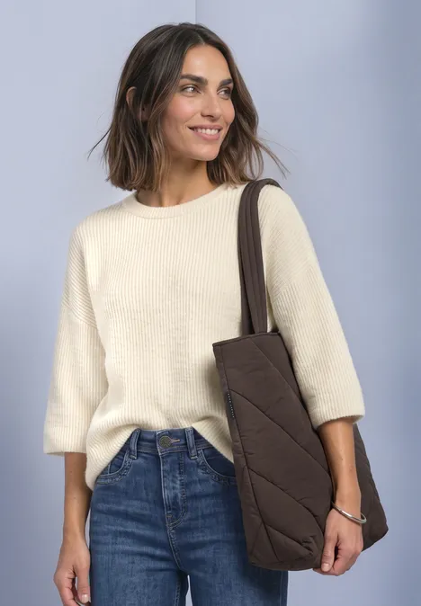 Sac cabas doublé, couleur unie auburn brown Frau mit cremefarbenem Pullover, blauen Jeans und brauner Stepp-Tasche, lächelnd vor neutralem Hintergrund.
