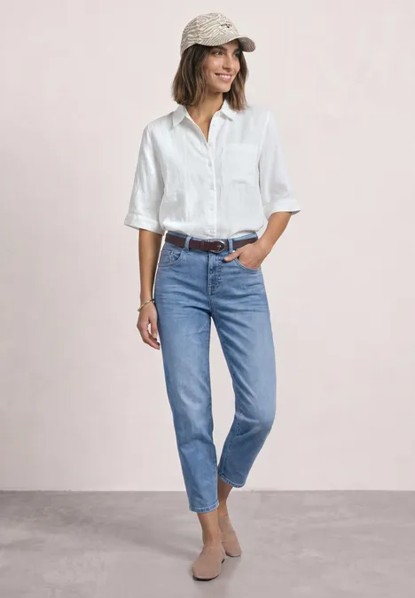 Frau in weißer Bluse, blauen Jeans, beigen Boots und Basecap steht entspannt vor hellem Hintergrund.