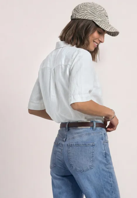 Frau mit weißem Hemd, blauen Jeans und gestreifter Cap steht seitlich und lächelt.