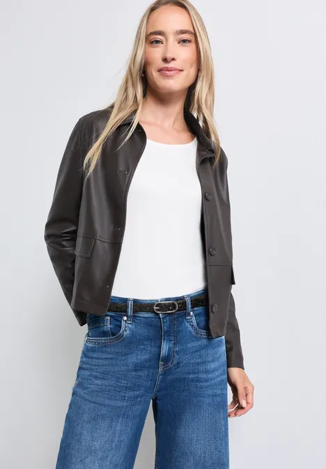 Eine Frau in weißem Top, schwarzer Lederjacke und blauen Jeans mit Gürtel lächelt.