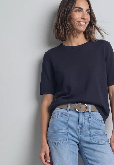 Riem met decoratieve gesp Muddy Mocha Frau lehnt lächelnd an grauer Wand, trägt schwarzes T-Shirt, helle Jeans und auffälligen Gürtel.
