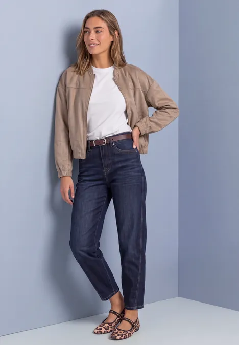 Eine junge Frau trägt eine beige Jacke, weißes T-Shirt, dunkle Jeans und gemusterte Sandalen vor hellblauem Hintergrund.