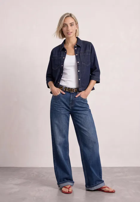Frau in Jeansjacke, weißem Top und weitem Jeans, orangenen Sandalen vor neutralem Hintergrund.