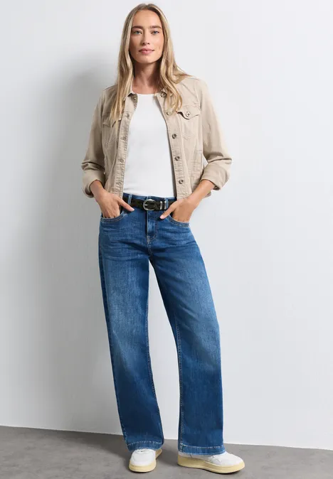 Frau trägt beige Jacke, weißes Shirt und weite blaue Jeans, steht entspannt vor neutralem Hintergrund.