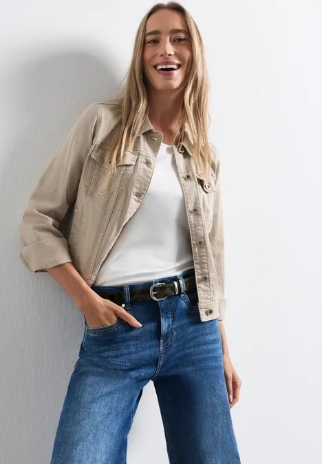 Eine lächelnde Frau in Jeans und heller Jacke vor weißem Hintergrund.