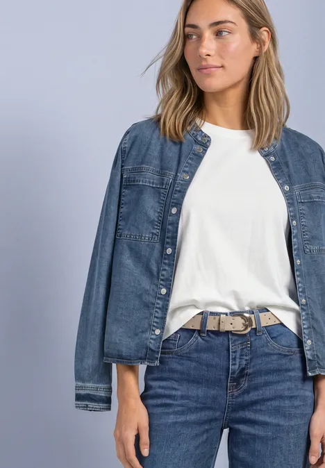Frau in weißem T-Shirt und Jeans mit lässiger Jeansjacke vor neutralem Hintergrund.