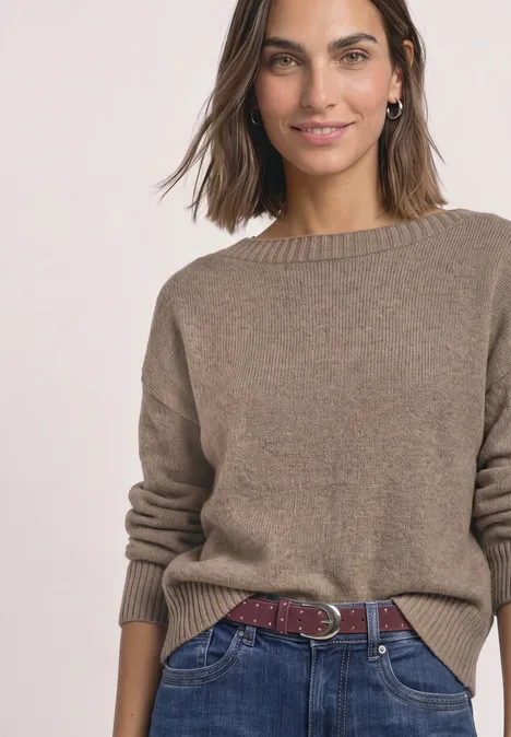 Frau in braunem Strickpullover, blauen Jeans und rotem Flechtgürtel, lächelnd.