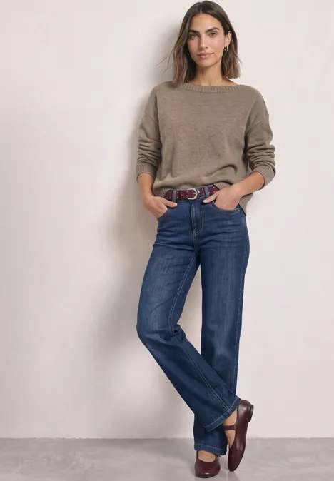 Eine Frau mit braunem Pullover, blauen Jeans und braunen Slippern lehnt entspannt an einer Wand.