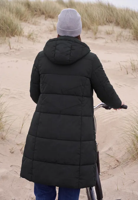 Steppmantel mit Kapuze und Reißverschluss Black Person im langen schwarzen Daunenmantel mit Mütze schiebt Fahrrad über sandige Dünen am Strand.