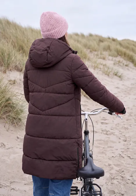 Steppmantel mit Kapuze und Reißverschluss berry red Person in langer brauner Steppjacke und rosa Mütze schiebt Fahrrad am Dünenstrand.