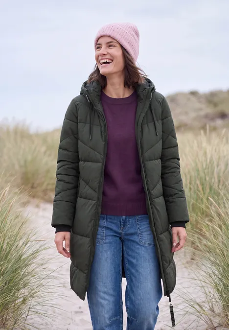 Steppmantel mit Kapuze und Reißverschluss olive night Eine lächelnde Frau in grüner Steppjacke mit Kapuze, rosa Mütze, Pullover und Jeans im Dünengras.