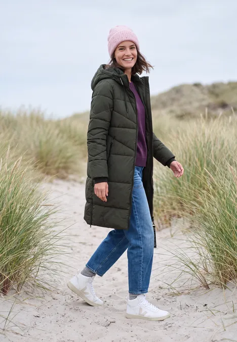 Steppmantel mit Kapuze und Reißverschluss olive night Frau im langen grünen Steppmantel, lila Pullover, Jeans und Beanie spaziert lächelnd über einen Dünenstrand.