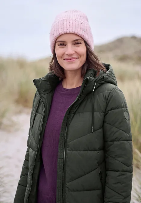 Steppmantel mit Kapuze und Reißverschluss olive night Eine lächelnde Frau am Strand, trägt eine rosa Mütze, lila Pullover und grüne Steppjacke.