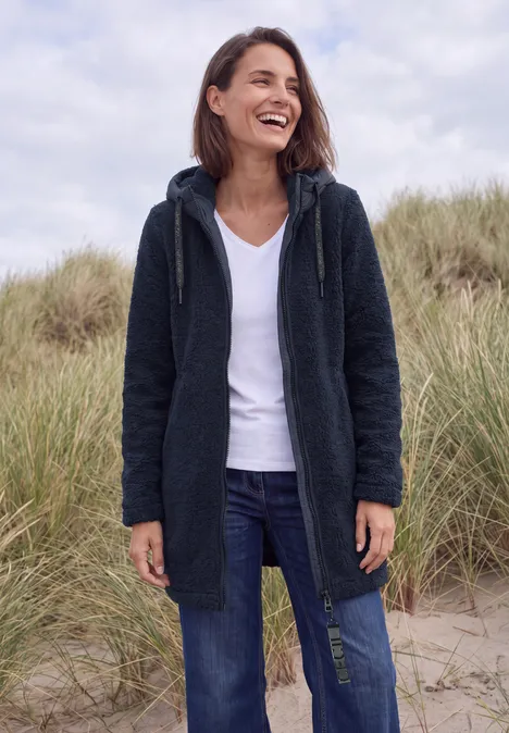 Strukturmix Teddy-Mantel mit Kapuze dark navy blue Lächelnde Frau in Kapuzenmantel und Jeans steht vor Dünen.