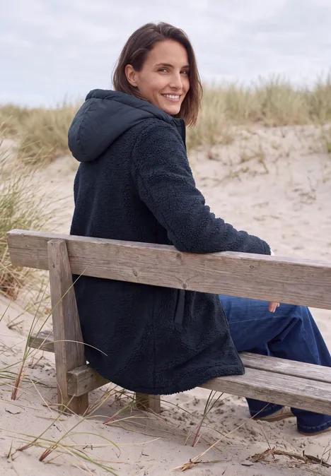 Strukturmix Teddy-Mantel mit Kapuze dark navy blue Eine lächelnde Frau sitzt auf einer Strandbank in den Dünen, trägt einen dunklen Mantel und schaut über die Schulter.