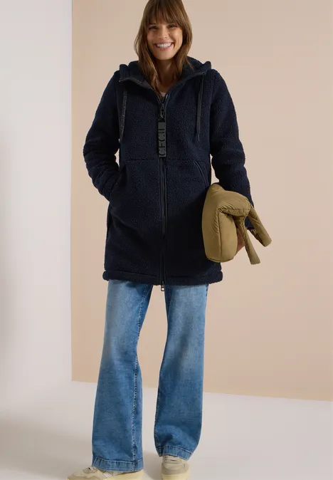 Manteau Teddy à texture contrastée avec capuche dark navy blue Frau im blauen Teddy-Mantel mit Kapuze, weiten Jeans und grünem Accessoire vor hellem Hintergrund.