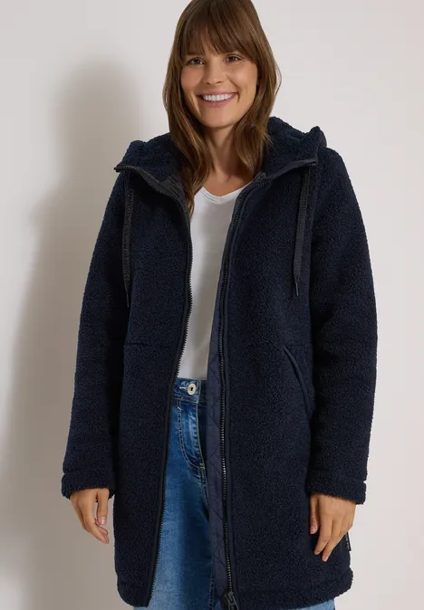 Manteau Teddy à texture contrastée avec capuche dark navy blue Frau in dunkelblauer, kuscheliger Kapuzenjacke, weißem T-Shirt und Jeans, lächelt entspannt vor hellem Hintergrund.