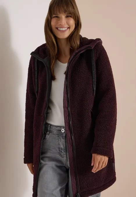 Frau mit dunkelrotem, kuscheligem Teddy-Mantel, Kapuze, weißem Shirt und Jeans, lächelnd vor hellem Hintergrund.