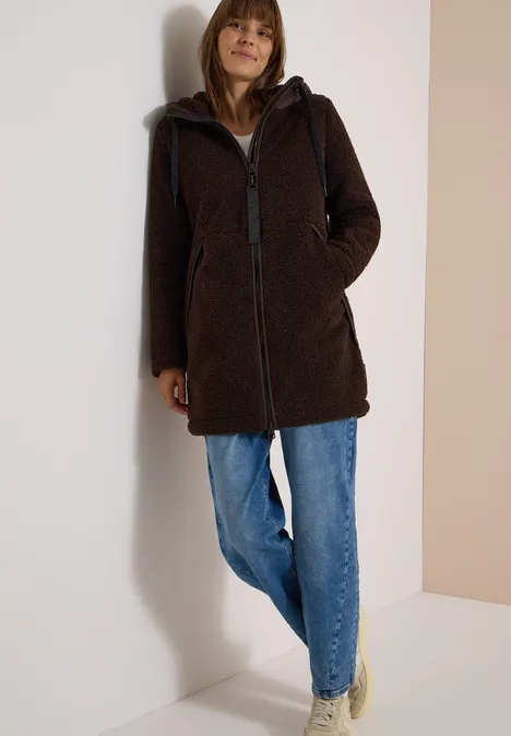 Strukturmix Teddy-Mantel mit Kapuze chocolate sauce brown Frau lehnt entspannt an Wand, trägt dunkelbraunen Teddy-Mantel mit Kapuze, Reißverschluss und Jeans.