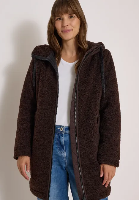 Strukturmix Teddy-Mantel mit Kapuze chocolate sauce brown Frau trägt langen, kuscheligen braunen Teddy-Mantel mit Kapuze und Reißverschluss, kombiniert mit Jeans und weißem Shirt.