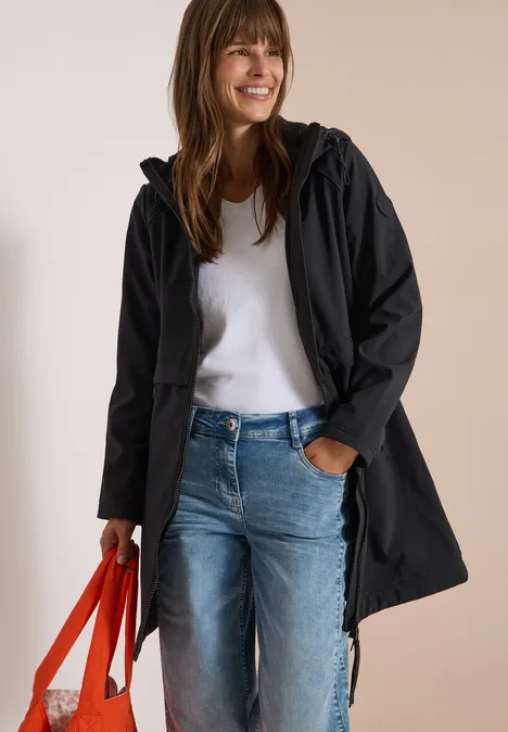 Frau lächelt in schwarzer Kapuzenjacke, weißem Pullover und Jeans, hält eine rote Einkaufstasche.