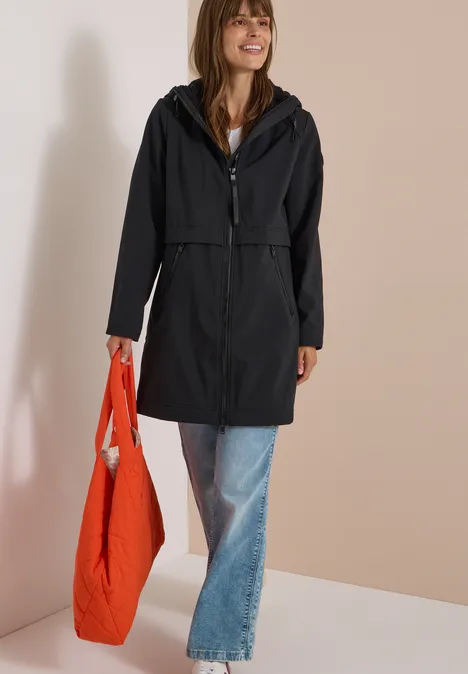 Frau in schwarzem Regenmantel und Jeans, lächelnd mit großer oranger Tasche vor hellem Hintergrund.