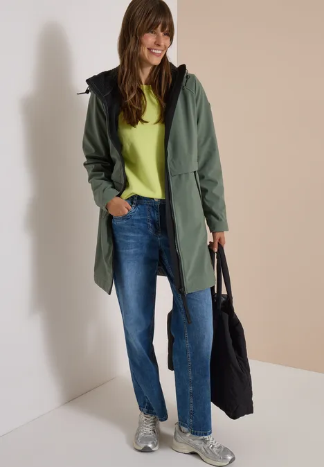 Une femme souriante en manteau vert, jean et baskets, tenant un sac noir.