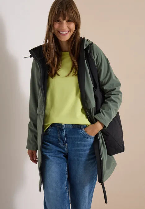 Une femme souriante porte un parka vert foncé, un t-shirt vert clair et un jean, sur fond clair.