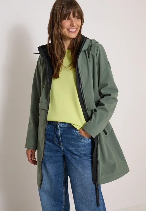 Femme souriante en imper vert à capuche, t-shirt jaune et jean bleu sur fond blanc.