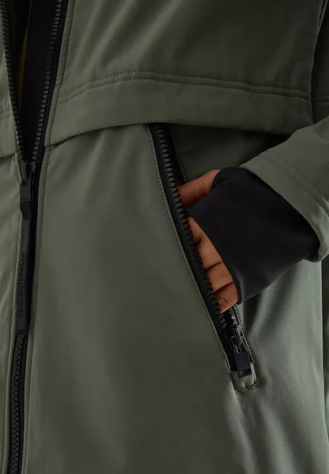 Une main dans la poche zippée d’une veste vert olive avec des poignets noirs.
